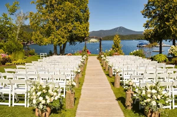 Allée de cérémonie en bois bordée de chaises blanches, face à une arche fleurie au bord d'un lac