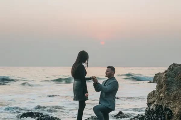 Un homme à genou tend la main à sa compagne sur des rochers en bord de mer, au coucher du soleil