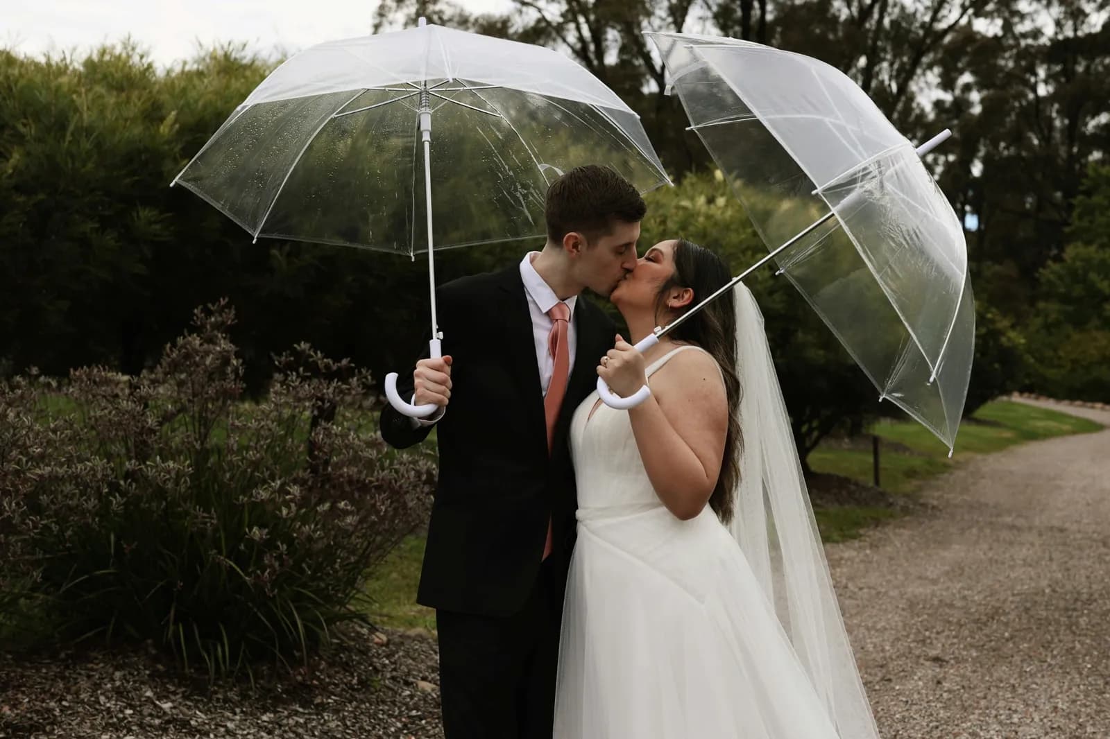 Jeunes mariés qui s'embrassent sous deux parapluies transparents dans un jardin